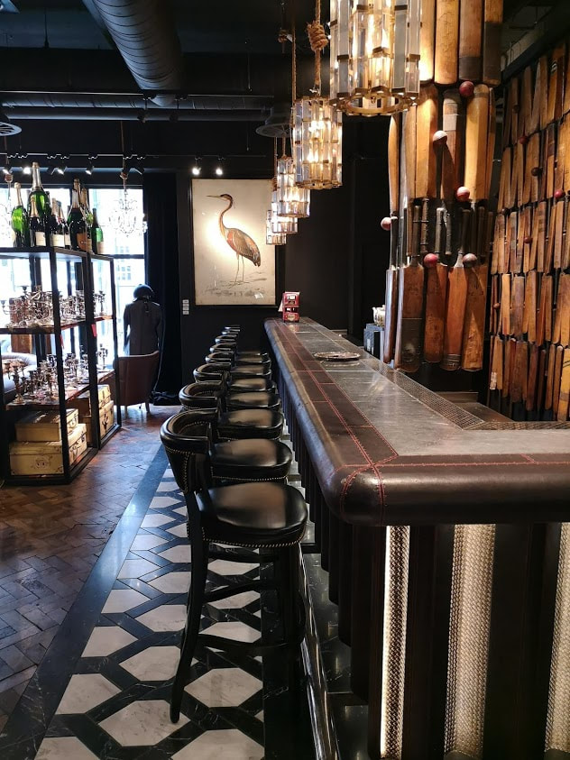 the bar at timothy oulton wwith cricket bats and balls as a backdrop and black leather stools in a row. 