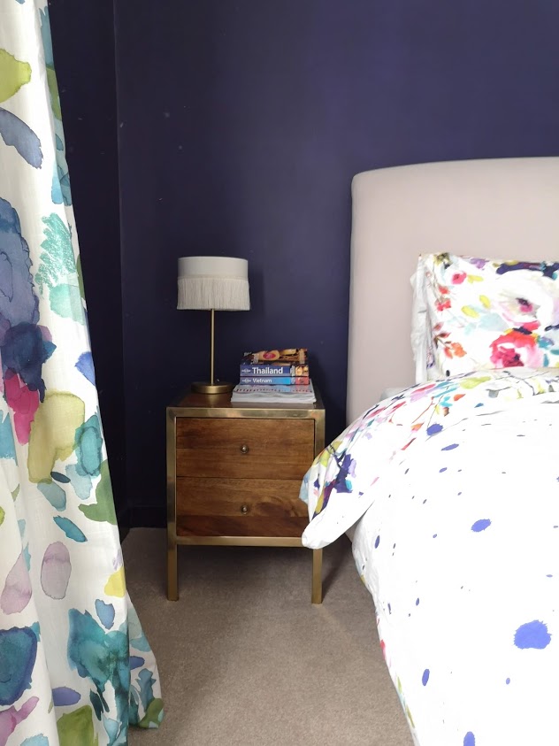indigo bedroom walls with abstract floral curtains and bedding. Brass bedside tables and fringed table lamp