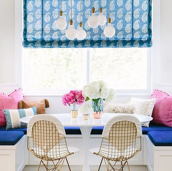 breakfast nook with cobalt blue seat coushins, white round marble table in the centre with three metallic vases with flowers in the centre. pink cushions on either corner are fringed with pompoms. There is a large bright window across the nook and a gold multi drop pendant above it. a roman blind hangs in the window with a paisley/leaf blue pattern on it. In front f the table are two gold wire framed chairs with fabric seat cushions 