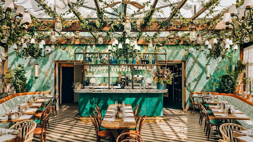 interiors photo of pink mamas in paris designed by Martin Brudnizki. the ceiling is covered with ivy and has multiple lights hanging from it. the walls are ina  distresed green finish with a bar in the centre with brass finishes. Around the perimeter of the room is a long booth with lots of small tables with bentwood chairs. 