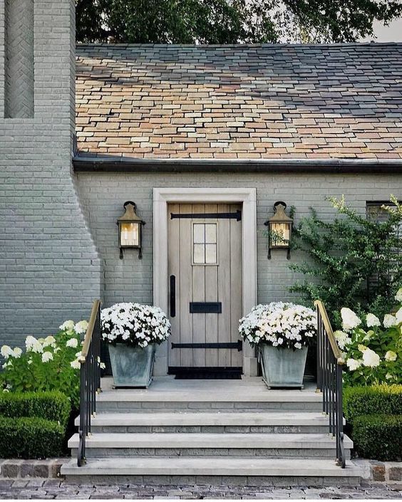 entrance doorway flanked by white hydrangeas and white flowers in zinc pots. the entrance is a few steps up from the ground level. the door is a rustic wood finish with black ironmongery. the door is flanked by two large lights.