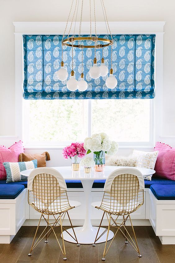 breakfast nook with cobalt blue seat coushins, white round marble table in the centre with three metallic vases with flowers in the centre. pink cushions on either corner are fringed with pompoms. There is a large bright window across the nook and a gold multi drop pendant above it. a roman blind hangs in the window with a paisley/leaf blue pattern on it. In front f the table are two gold wire framed chairs with fabric seat cushions 