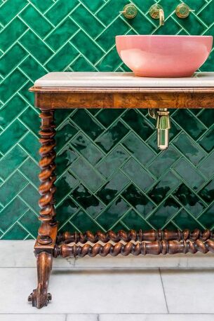 pink sink mounted on antique table with brass taps and gloss green tiles in a herring bone pattern across the wall. white marble floor tiles.