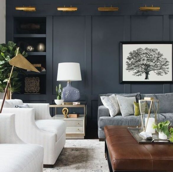 inky indigo panelled walls and brass lights above with a grey sofa on the right side. two cream arm chairs on the left and a leather ottoman in the centre. on the ottoman is a brass tray with some beautiful objects. in the corner is a large fiddle leaf fig tree. 
