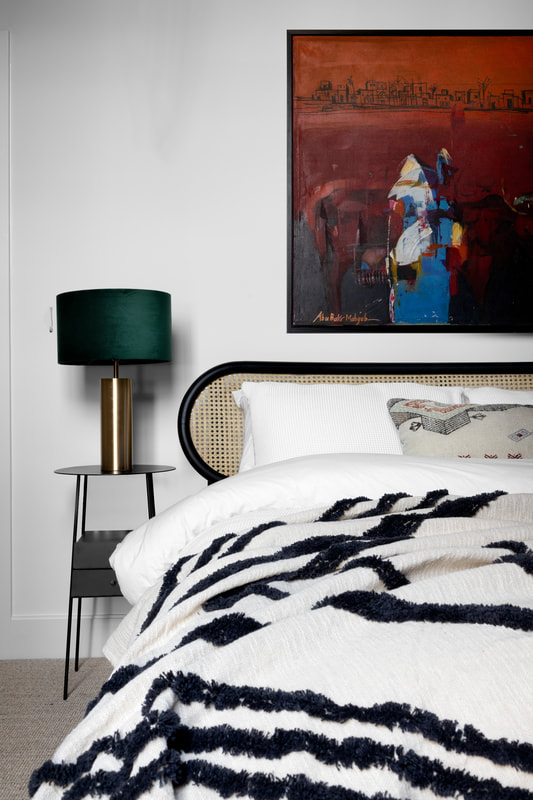 Vignette of bedroom with black metal bedside table and gold and green table light. Rattan headboard with textured patterned bedding. Dark red artwork above the bed framed in black.