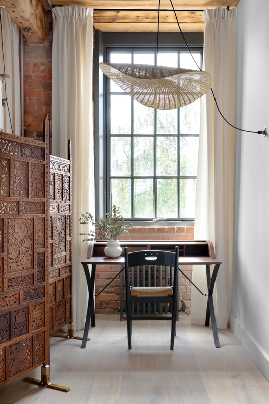 Writing desk facing out of a window flanked by long white curtains with a hand carved indian screen to the left. An interesting large rattan lampshade is suspended above the desk.