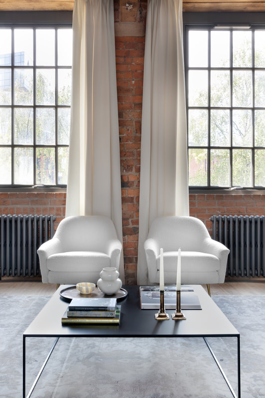 white boucle armchairs set against an exposed rustic red brick wall with green trees outside. White linen curtains